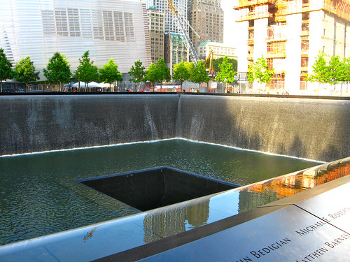 A large square recessed pool with a smaller square hole in the center. Behind the pool are some trees, and behind those are some buildings, some of which are under construction. In the foreground, we see the rim of the pool lists names.