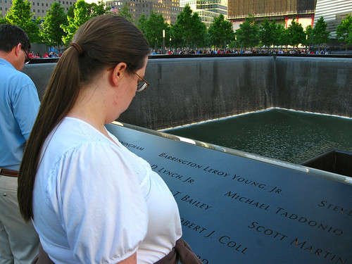 Sarah, a woman in her early thirties with long, brown hair in a ponytail, wearing a white blouse, facing away from the camera, solemnly contemplates the names on the rim of a recessed pool. There are trees on the other side of the pool, and tall buildings on behind the trees.