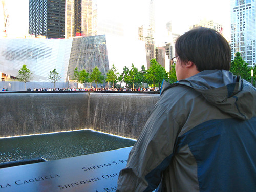 Chris, a man in his early thirties with short dark brown hair, wearing a gray and blue windbreaker, looks over the rim of a recessed pool. Names are visible on the rim of the pool. Behind the pool are trees, and behind the trees are tall buildings, and a shorter glass building.