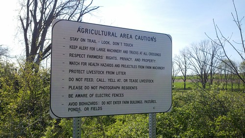 A white sign is posted in front of some bushes, behind which is a farm. The sign reads: Agricultural area cautions: Stay on trail. Look. Don't touch. Respect farmer's rights, privacy, and property. Watch for health hazards and projectiles from farm machinery. Protect livestock from litter. Do not feed, call, yell at, or tease livestock. Please do not photograph residents. Be aware of electric fences. Avoid biohazards: Do not enter farm buildings, pastures, ponds, or fields.