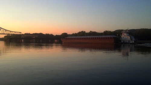 A barge moves up a river at sunset. The sunset is reflected in the river. On the other side of the channel are a silhouette of trees. On the left of the photo, a silhouette of part of a cantilever bridge is seen.