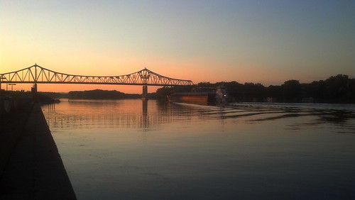 A barge moves up a river at sunset. The sunset is reflected in the river. On the other side of the channel are a silhouette of trees. We also see the silhouette of most of a cantilever bridge, and the walkway on which the photograph was taken. The barge is almost under the bridge.