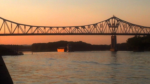 A cantilever bridge crosses a river. Behind it is a barge upstream and some hills. The sky is a gradient from yellow to deep orange, some of which is reflected in the river.