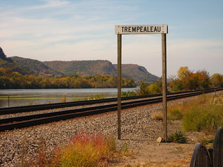 A pair of train tracks along the Mississippi river. Next to the tracks in a railroad sign reading "Trempealaeu".