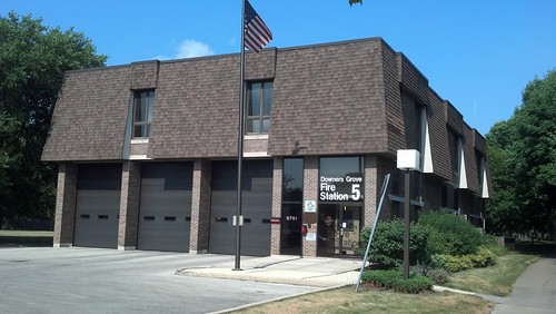 Downers Grove Fire Station 5: A two-story fire station with three garage doors and a glass window with door in its front. The name of the station is printed on the glass. There is a flagpole with a U.S. flag in front as well.