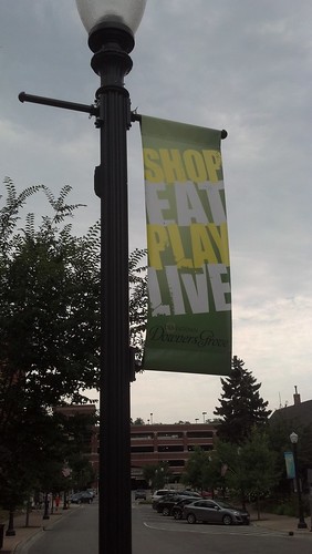 A lamp-post with a vertical banner reading "Shop, Eat, Play, Live". The words are alternating yellow and white, and the background is green.