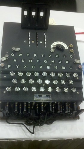 An Enigma machine: It's a black box, roughly the size of a suitcase. On the top of the machine is a typewriter-style keyboard in QWERTZ layout. Behind the keyboard and another set of lights labelled with letters in a QWERTZ layout, and behind that are a set of three rotors. On the front are a bunch of wires and plugs.