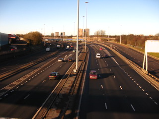 A view of a British motorway (what I would call a freeway in American English) from a pedestrian bridge. It's a divided highway with two lanes on the left of traffic moving away from us and three lanes on the right with traffic moving toward us. There are on-ramps merging in both directions. In the center is a median lined with tall streetlights. In the distance are several tall buildings.