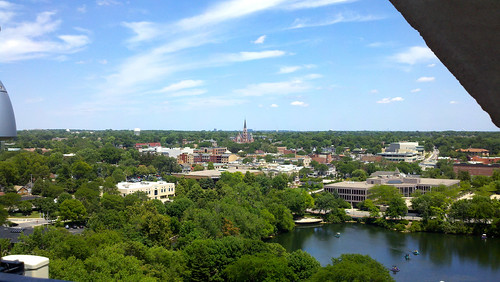 An aerial shot of downtown Naperville, Illinois from the top of the Millennium Carillon. There is a lake surrounded by short buildings, poking through a blanked to green trees. The horizon is flat, but if you look very closely, there are bits of very distant Chicago skyscrapers visible.