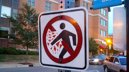 A hand painted regulatory traffic sign, in which a pedestrian figure is behind a red no-slash, but has broken part of the slash with their arm. There are red triangles emanating from where the figure's arm has broken the slash, representing shattered shards of the slash.