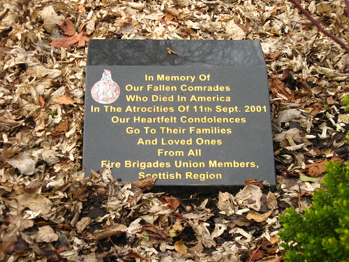 A small stone memorial reading "In Memory Of Our Fallen Comrades Who Died in America In The Atrocities Of 11th Sept. 2001. Our Heartfelt Condolences Go To Their Families And Loved Ones. From All Fire Brigades Union Members, Scottish Region." The memorial is surrounded by brown leaves.