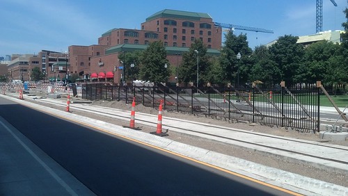 An not yet built light rail station in the median of a city street. There are a few traffic cones along the edge of the median. In the background is are some large buildings, some trees, and some construction cranes.