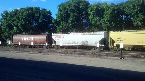A connected trio of rail cars sits on a track on the far side of a parking lot. One sports the "Canada" wordmark while another is labeled "CP Rail". Behind the cars are some tall trees.